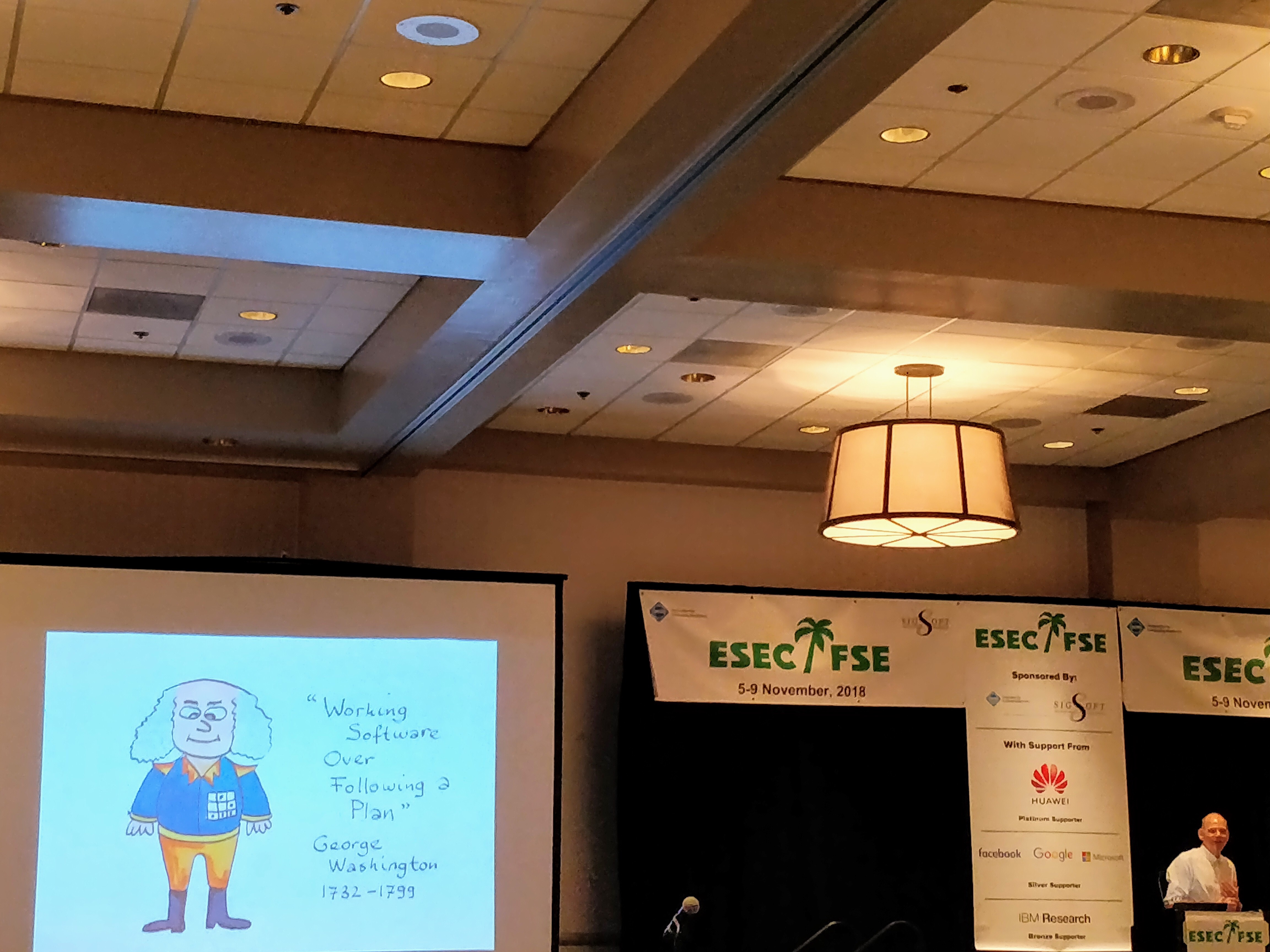 A conference-room view at ESEC/FSE 2018, with a joke slide showing a cartoon George Washington captioned "Working Software Over Following a Plan" and the ESEC/FSE 2018 banner in the background